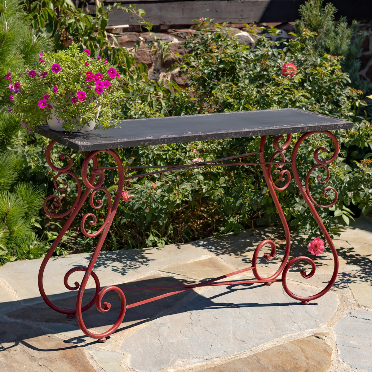 Rectangular dark tabletop on ornate red scrollwork legs, displayed outdoors with a potted flowering plant.