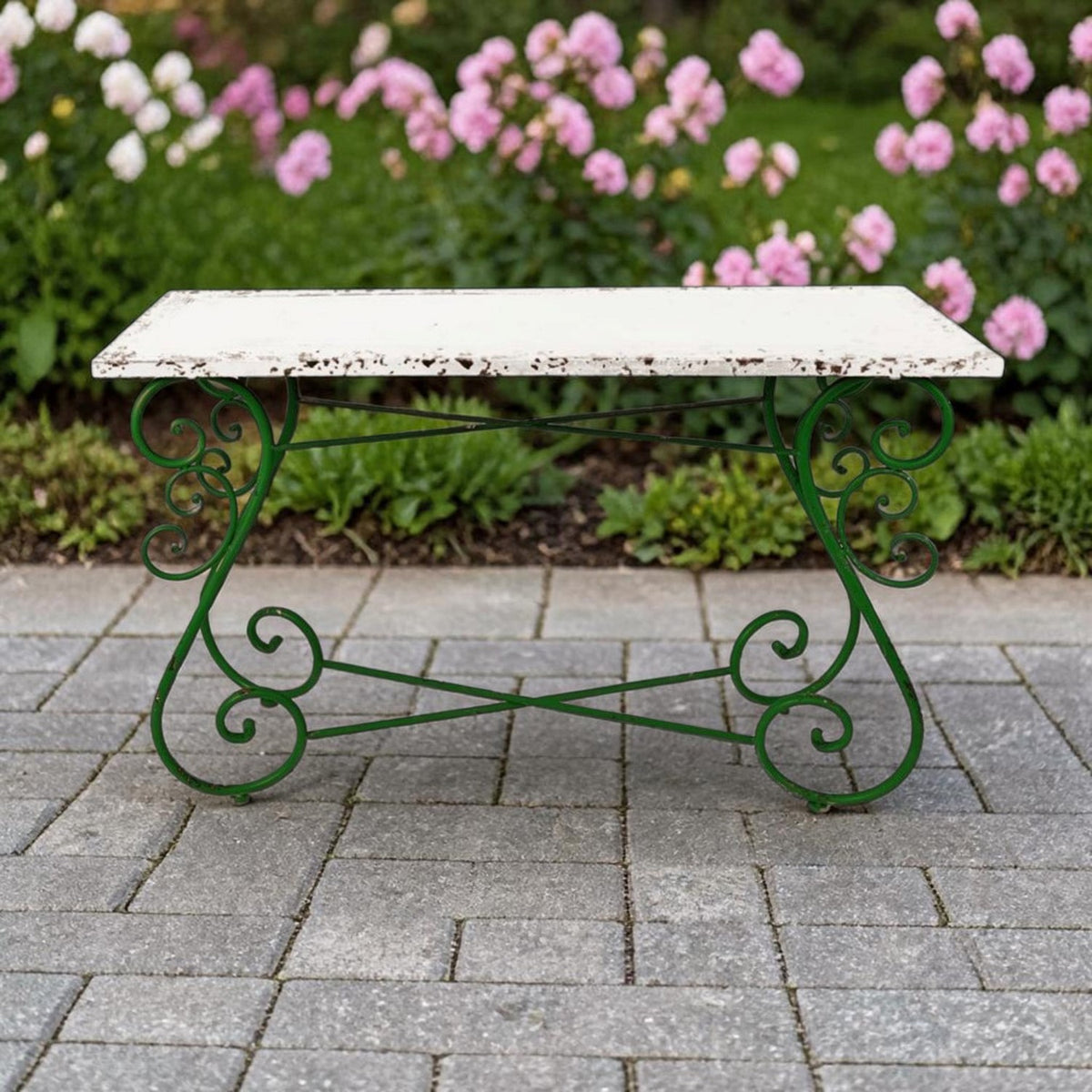Front view of a pale, distressed rectangular tabletop on dark green curled metal legs against a garden backdrop.
