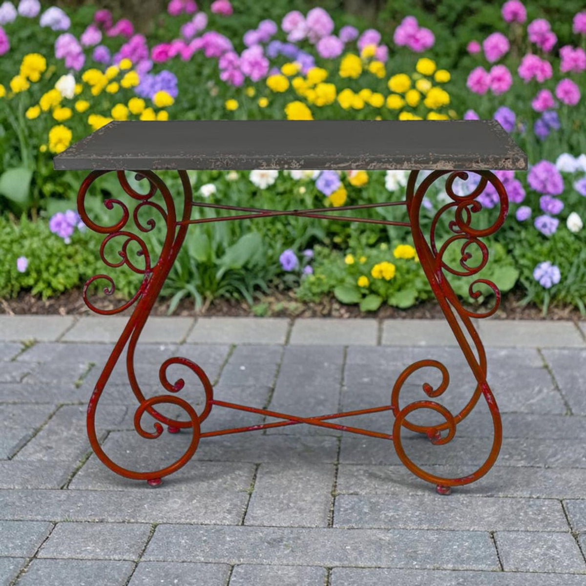 Dark rectangular tabletop on an ornate reddish iron frame, shown on a paved patio beside colorful flowers.