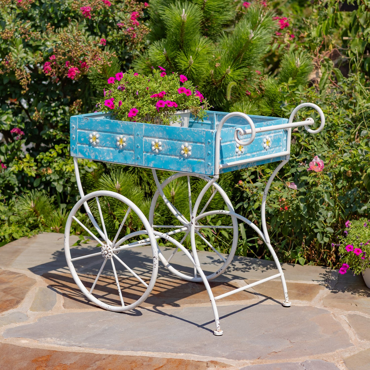 Blue flower cart filled with pink blooms, shown on a stone path among garden greenery.