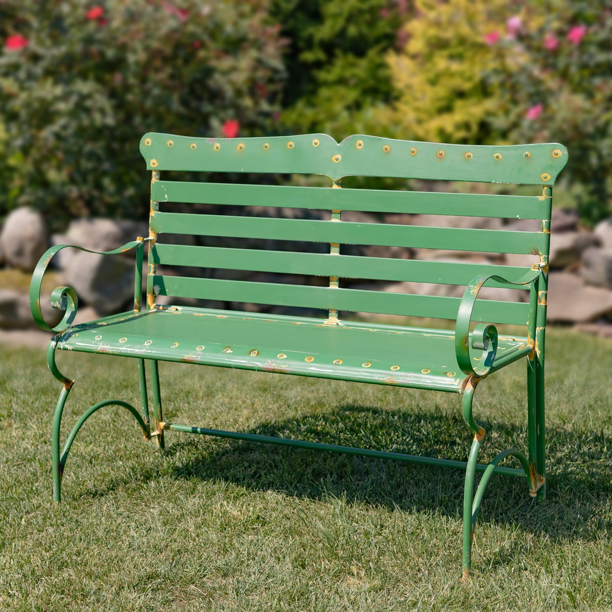 Green metal garden bench with slatted back and curled arms on a lawn among flowering shrubs.