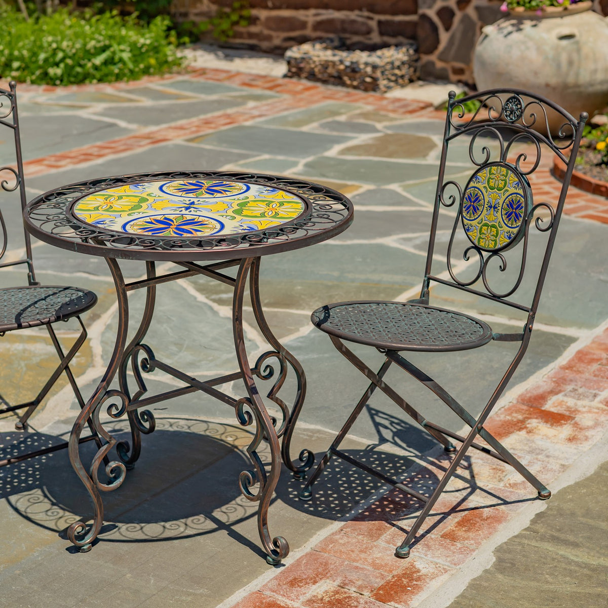 Round mosaic bistro table beside two ornate folding chairs on a stone patio.