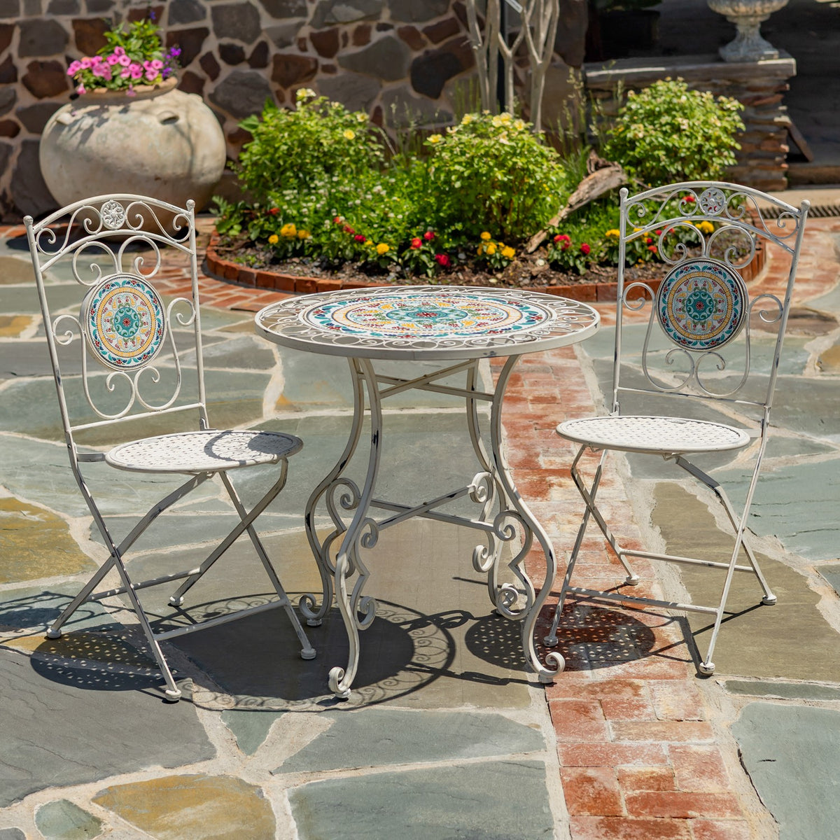Round mosaic bistro table with two ornate white metal folding chairs on a stone patio.