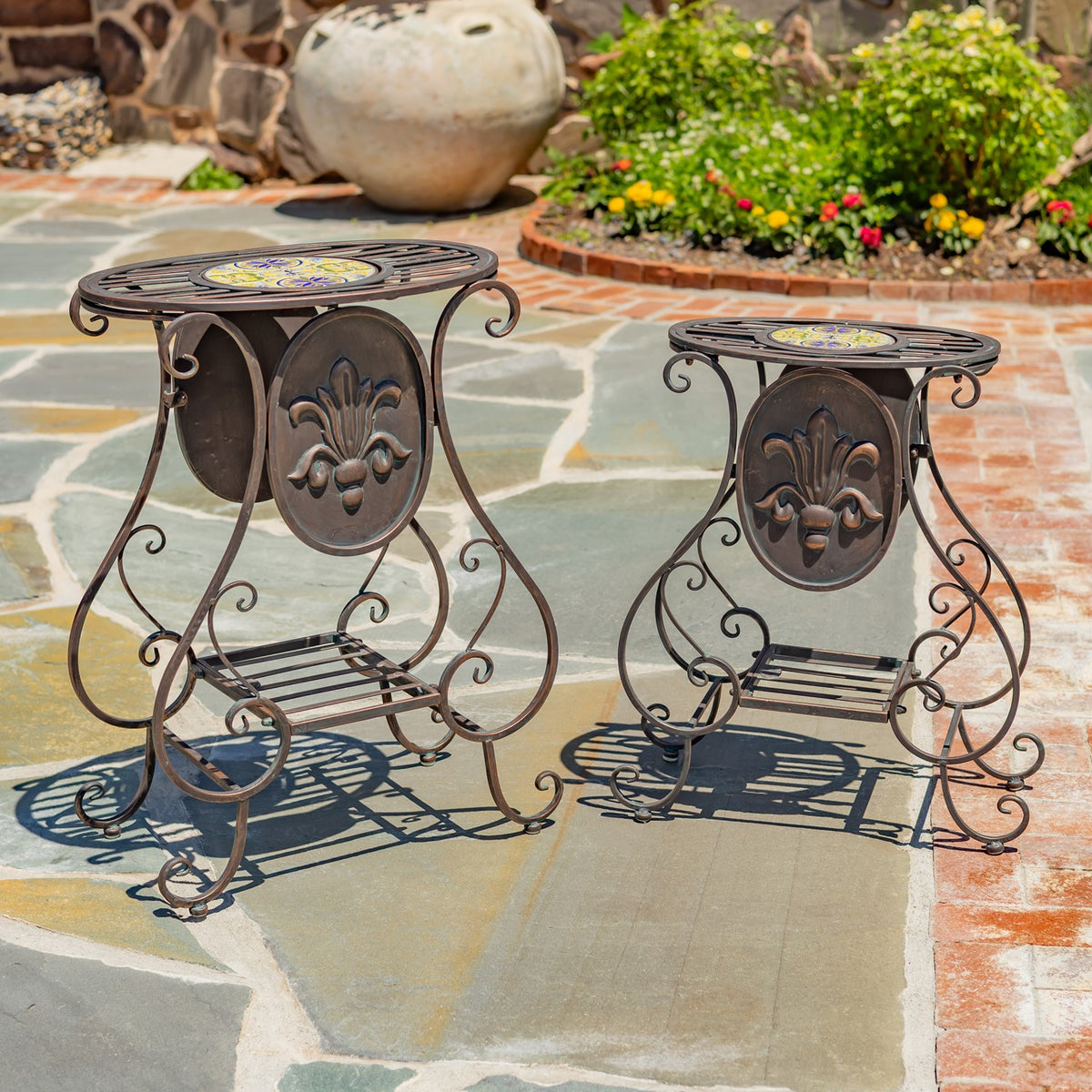 Two ornate metal plant stands with round mosaic tops displayed on a stone patio.