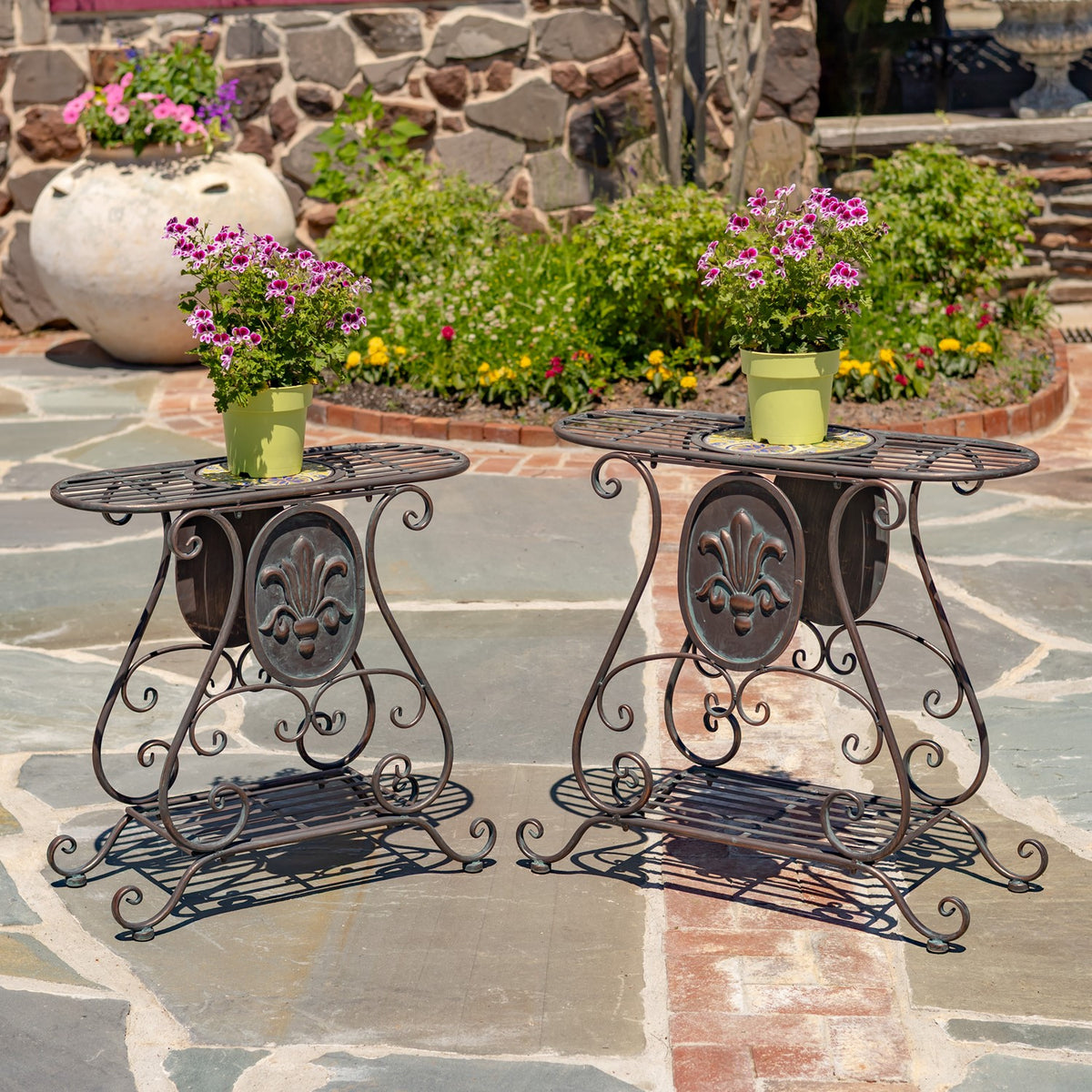 Two ornate dark metal plant stands with potted flowers on a stone patio.