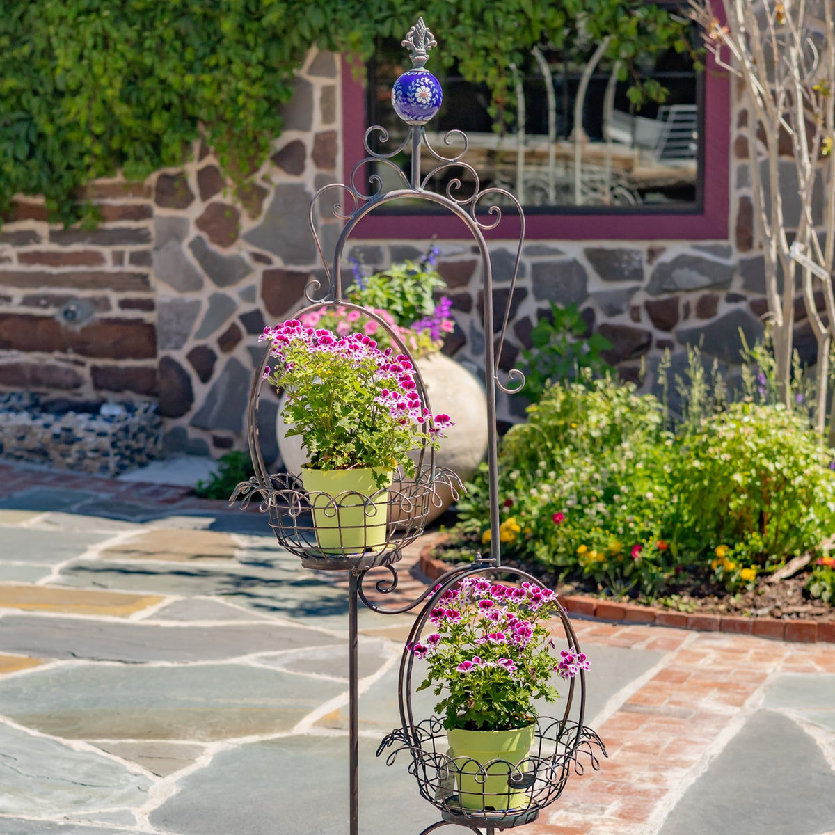 Ornate black wire plant stand with two flower-filled baskets on a stone patio