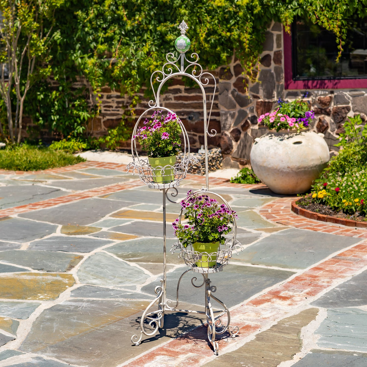Ornate white metal plant stand with two flower-filled wire baskets on a stone patio.