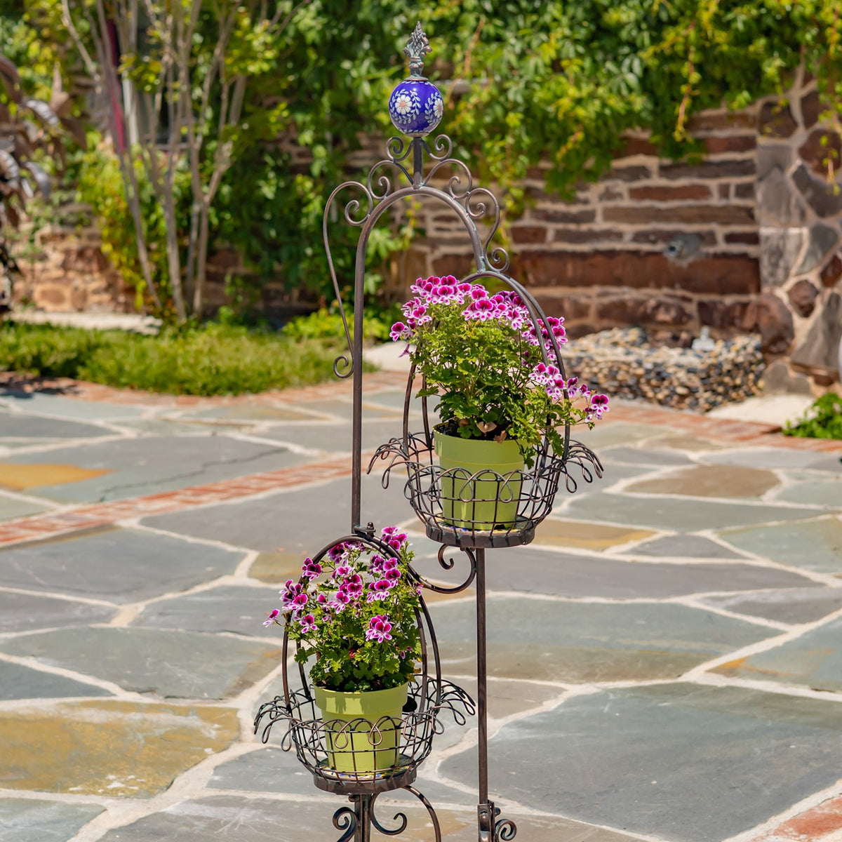 Ornate bronze plant stand with two flowering plants in wire baskets on a stone patio.