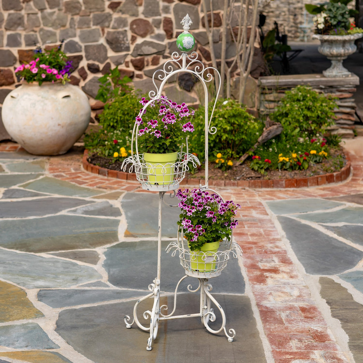 Antique white scrollwork plant stand with two flower-filled wire baskets on a stone patio.