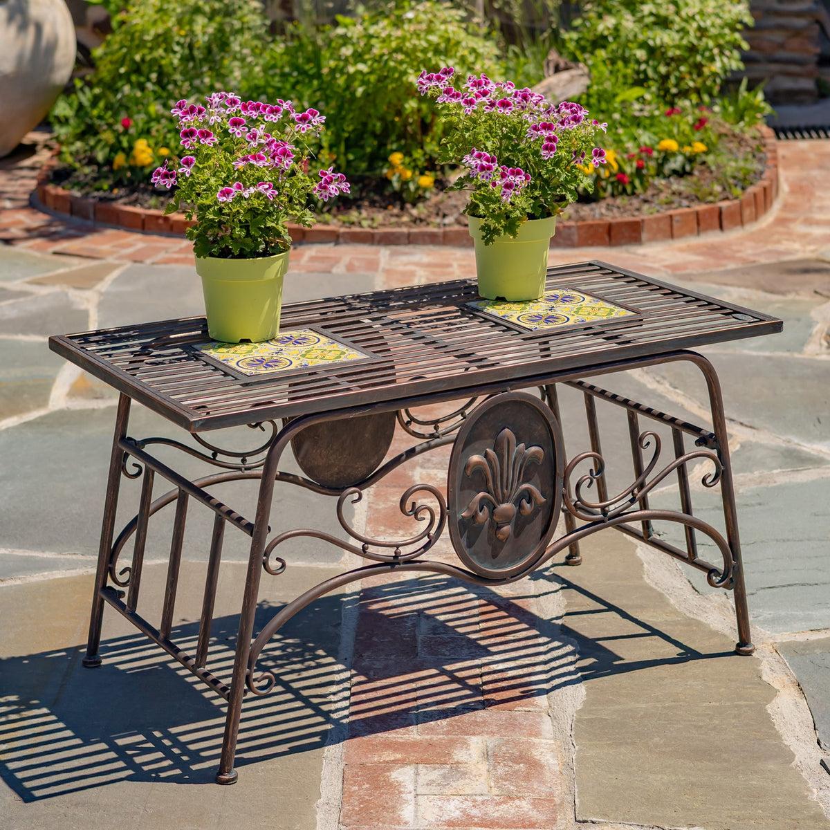 Ornate rectangular metal table with colorful mosaic insets and two flower pots on a stone patio.