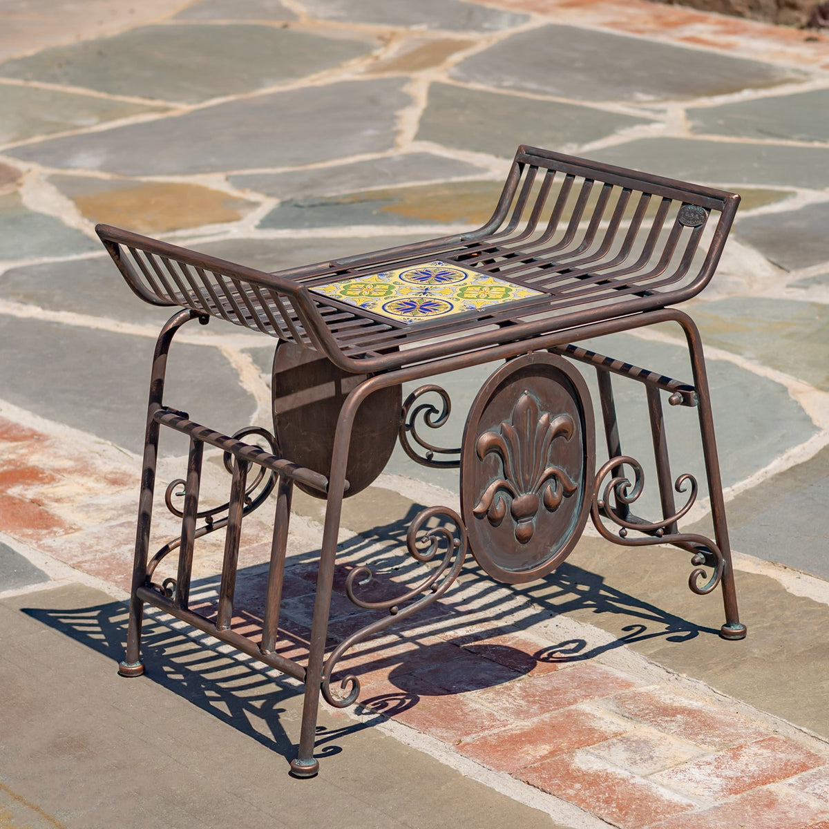 Backless metal chair with a colorful mosaic seat and ornate scrollwork, shown at an angle on stone paving.