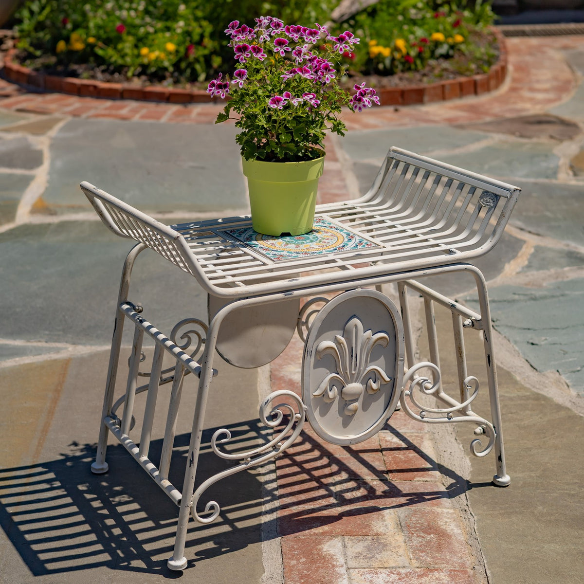 White metal chair with a colorful mosaic seat panel and ornamental side scrollwork, shown outdoors with a potted plant.