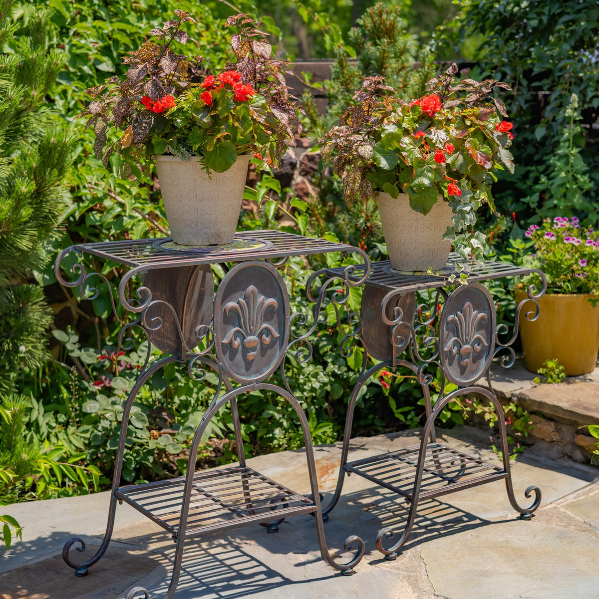 Two ornate metal plant stands on a stone patio, each topped with a flowering potted plant.