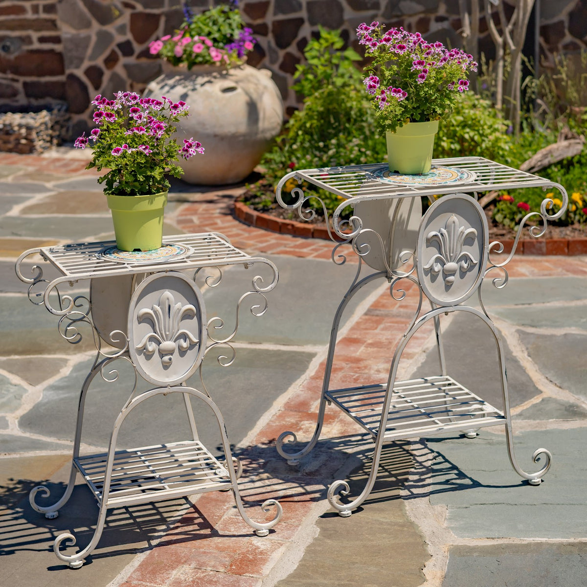 Two ornate metal plant stands with pink flowers on a stone patio.