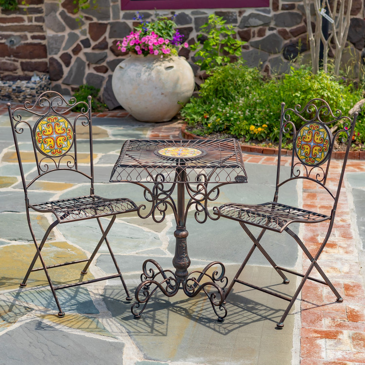 Ornate metal bistro table and two folding chairs with colorful mosaic accents on a stone patio.