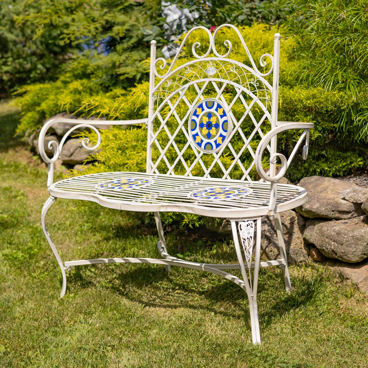 White metal garden bench with blue and yellow mosaic accents among shrubs and grass.