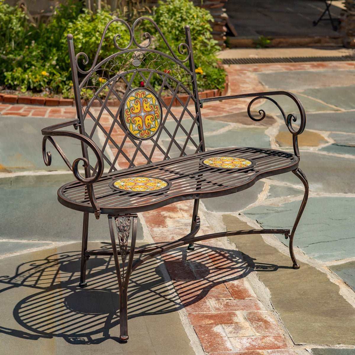 Ornate dark metal garden bench with colorful mosaic inlays on a stone patio, viewed at an angle.