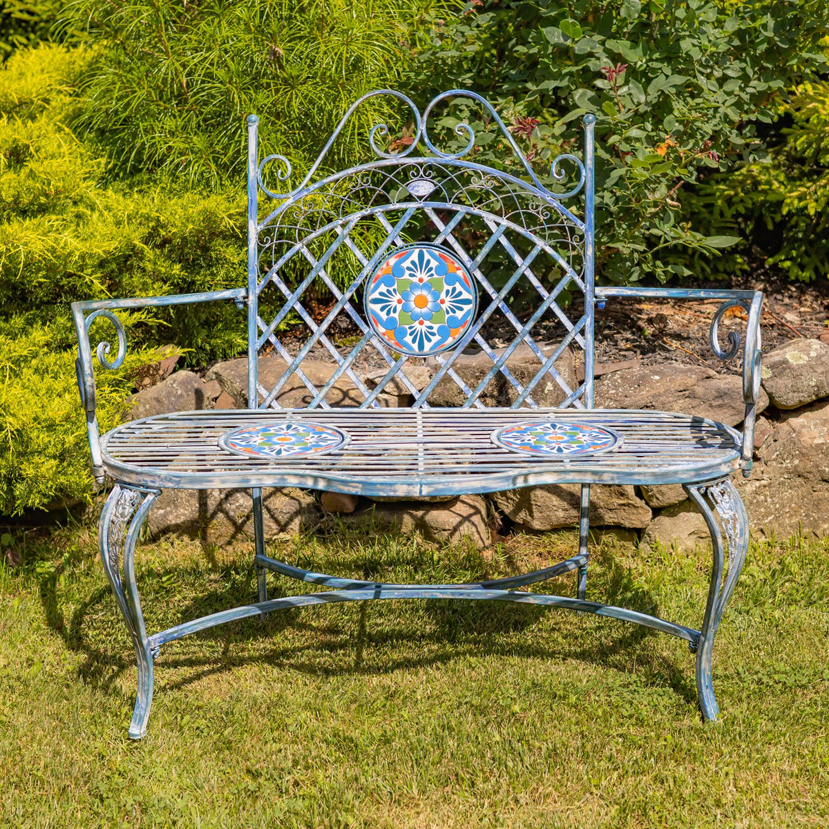 Ornate metal bench with colorful mosaic accents on a lawn among garden plants.
