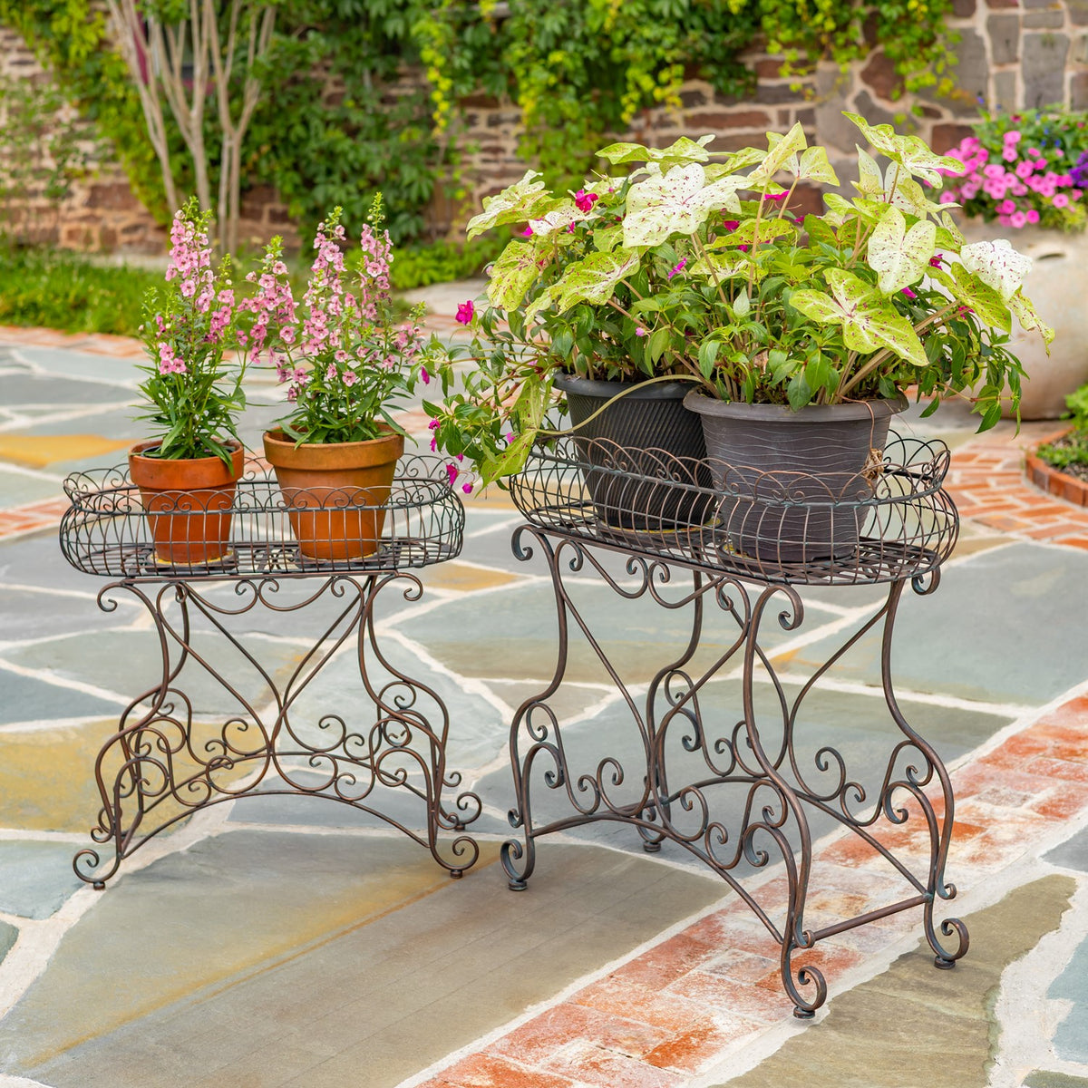 Two ornate bronze-toned metal plant stands display potted flowers and foliage on a stone patio.