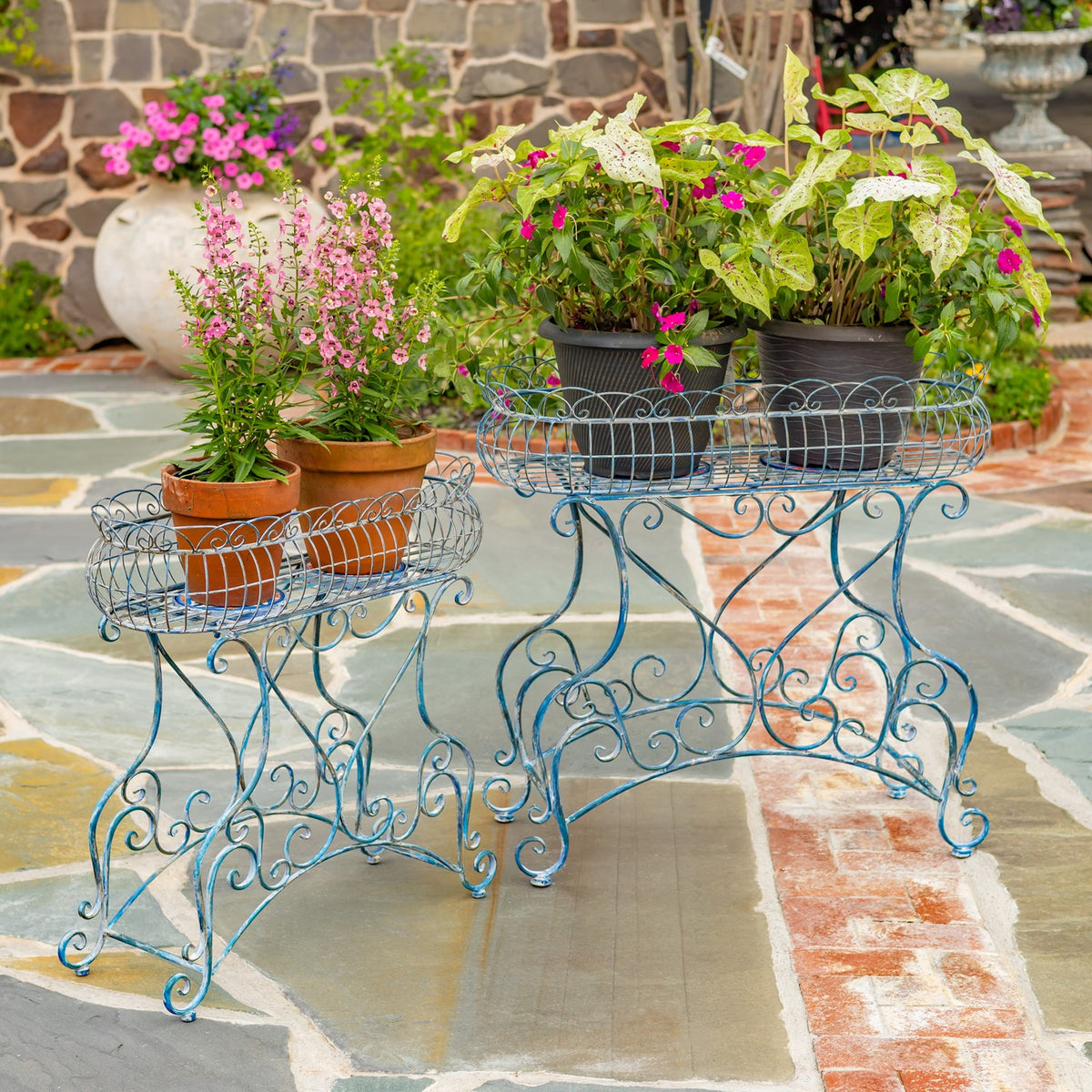 Two ornate blue metal plant stands hold potted flowers on a stone patio.