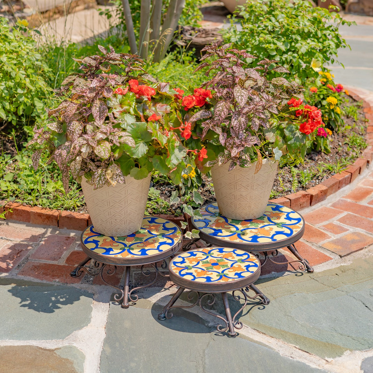 Three colorful mosaic-topped plant stands arranged in a garden, with flowering pots on the two taller stands.