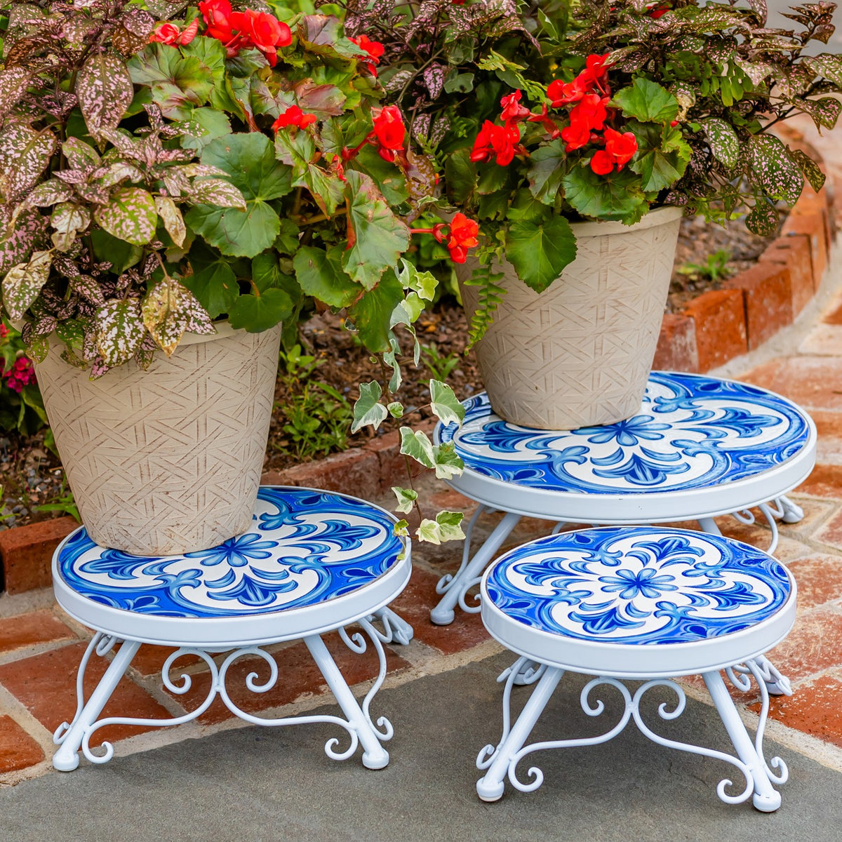 Three white metal plant stands with blue patterned tops arranged beneath potted flowers.