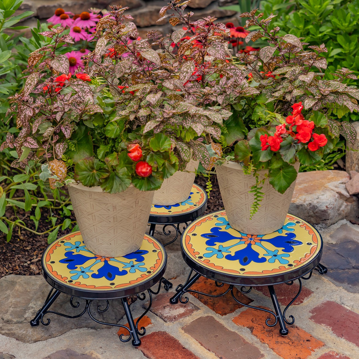 Two colorful mosaic plant stands hold potted flowers in a stone garden; a third stand peeks out between them.