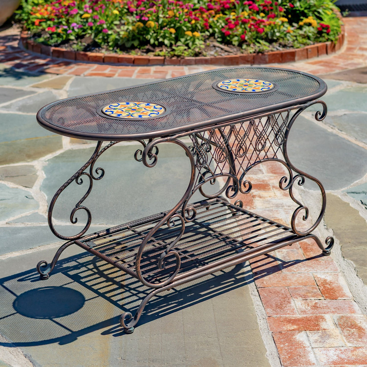 Ornate oval metal garden table with two colorful mosaic insets and a slatted lower shelf on a stone patio.