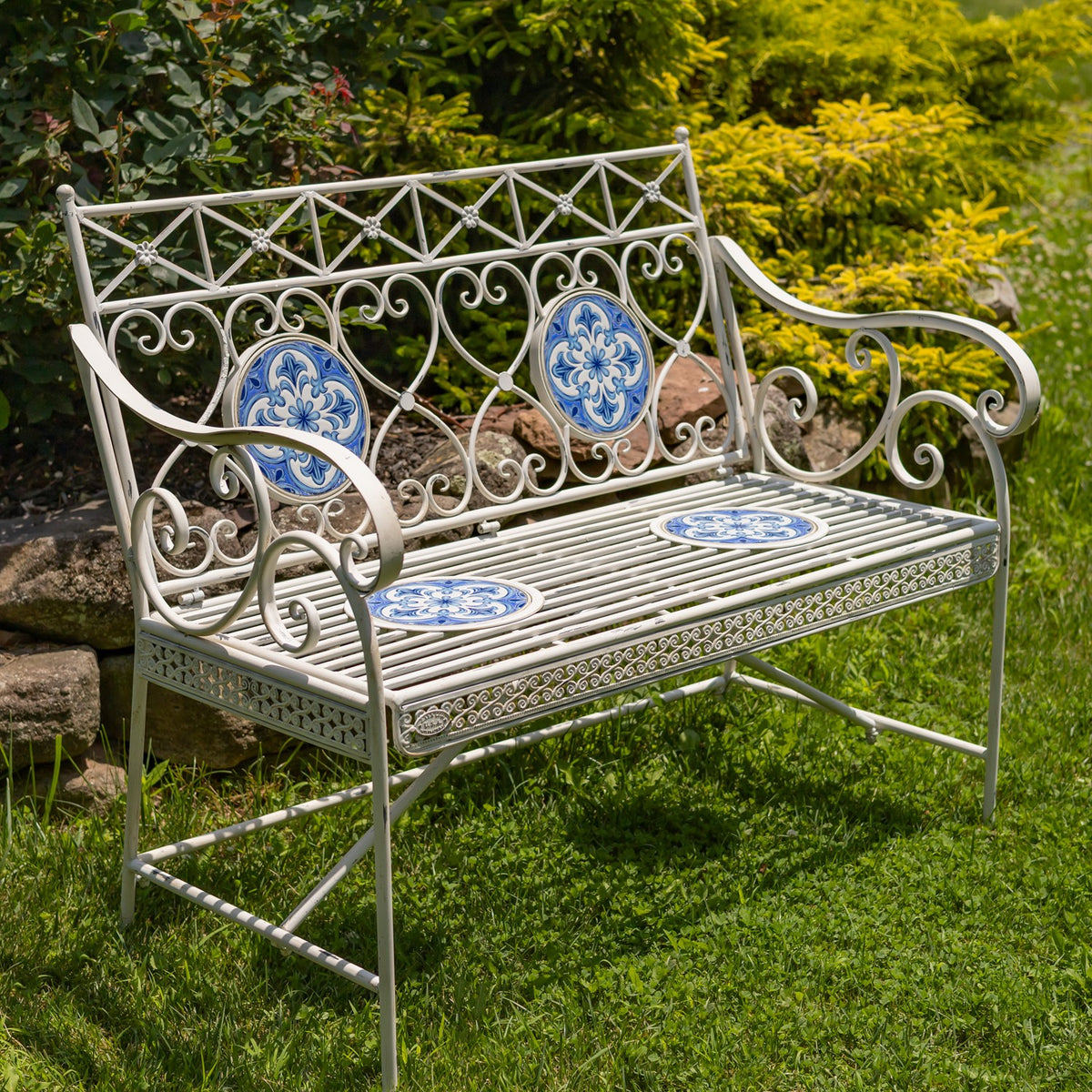 Antique-white metal garden bench with blue patterned mosaic inserts, set on grass among leafy shrubs.