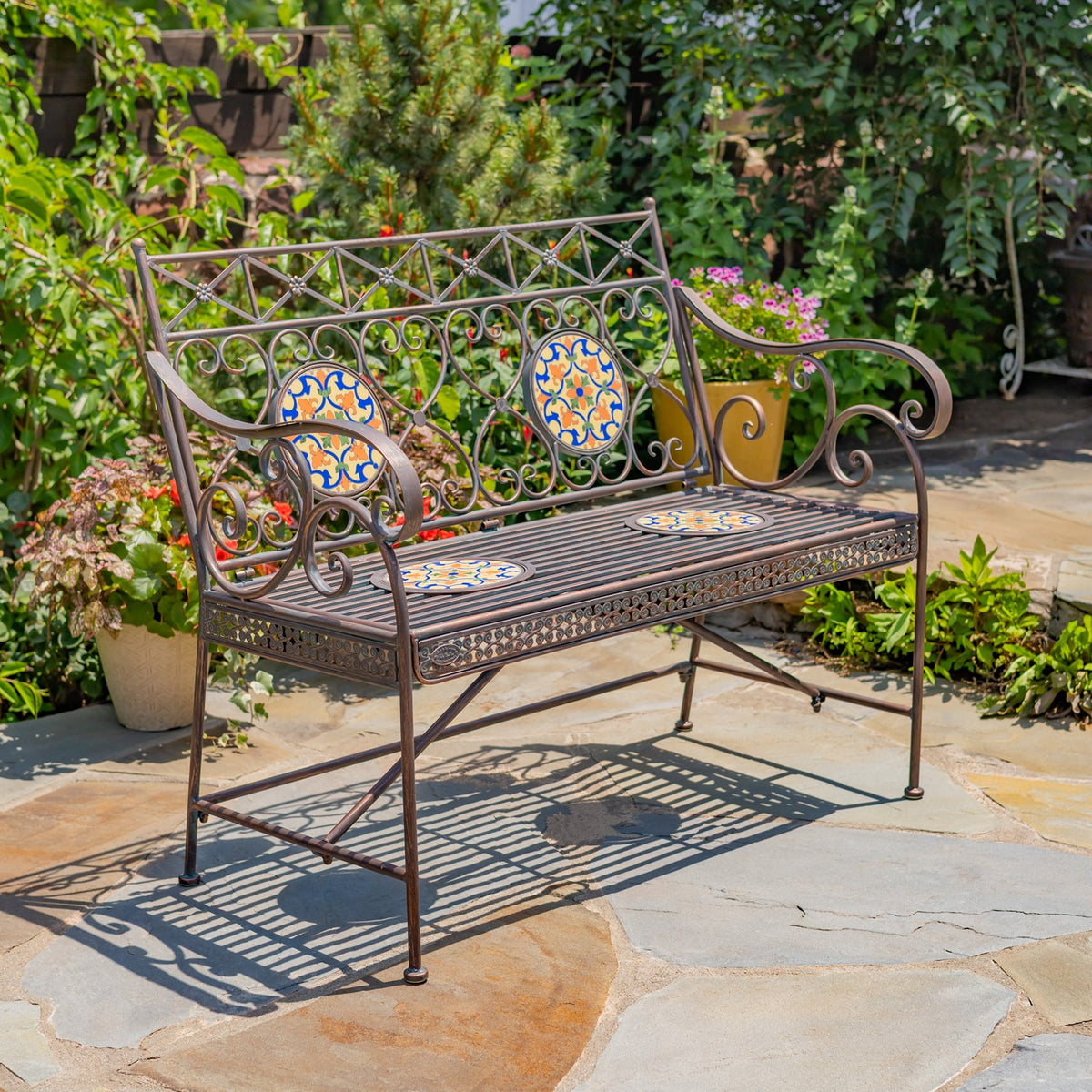 Ornate bronze-toned metal bench with colorful mosaic insets on a stone patio among greenery.