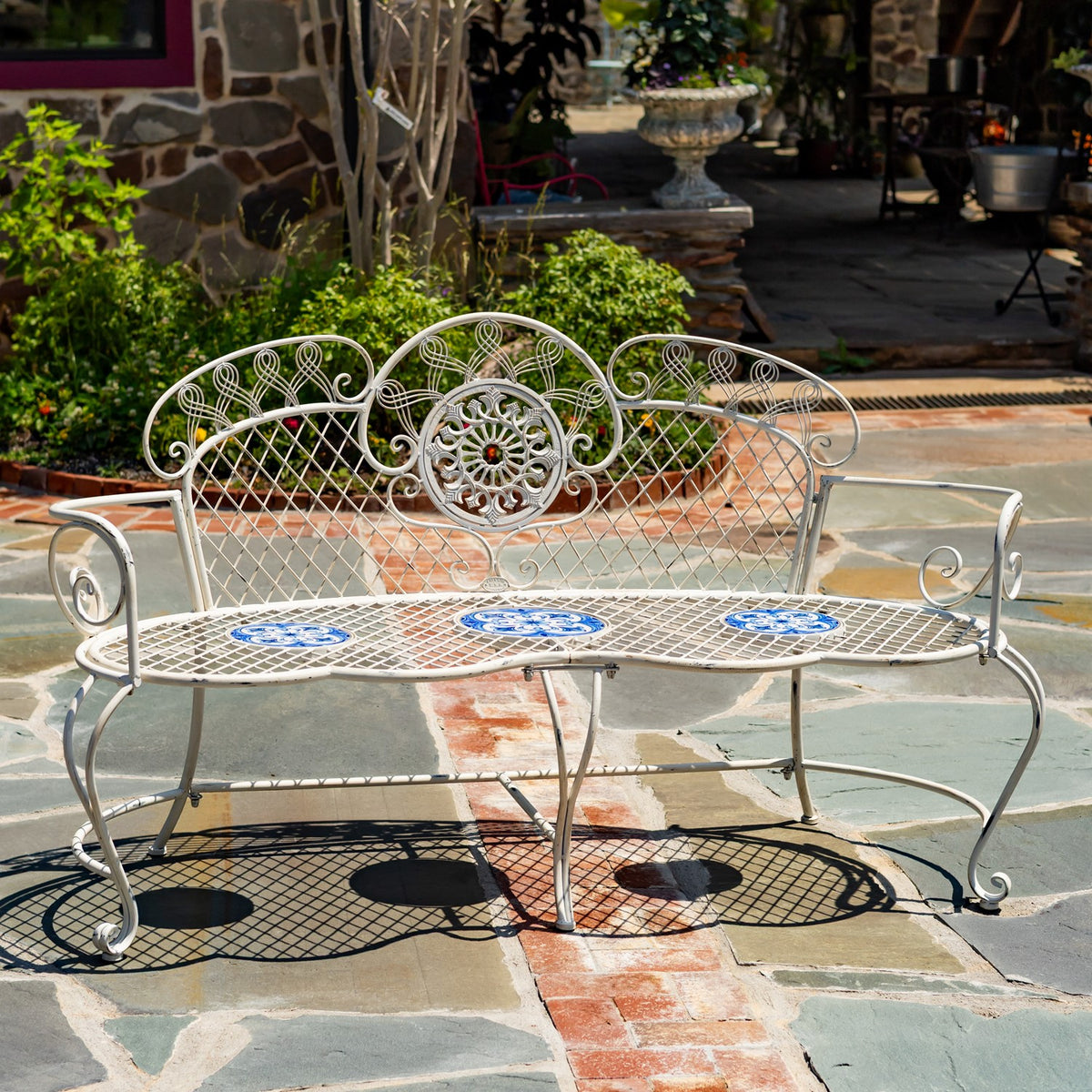 Ornate pale metal garden bench with blue mosaic seat accents on a stone patio.