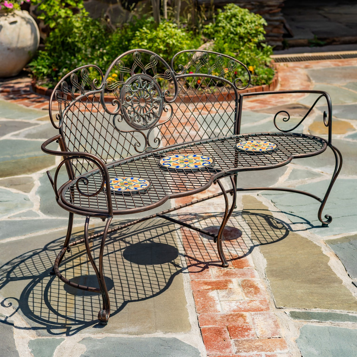 Ornate dark metal garden bench with three colorful mosaic seat tiles on a stone patio.