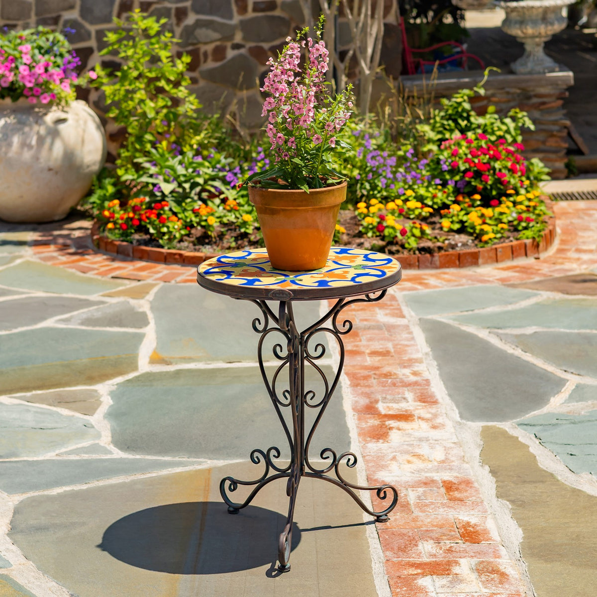 Mosaic-topped plant stand holding a terracotta pot with pink flowers in a sunny garden patio.