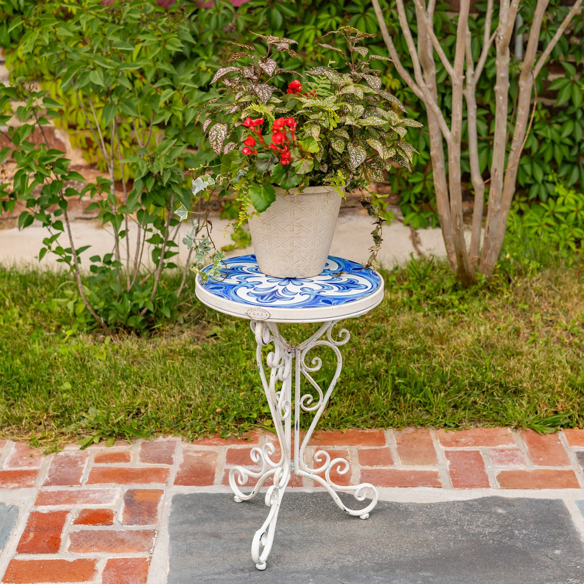 White wrought-iron plant stand with a blue-and-white mosaic top holding a potted flowering plant outdoors.