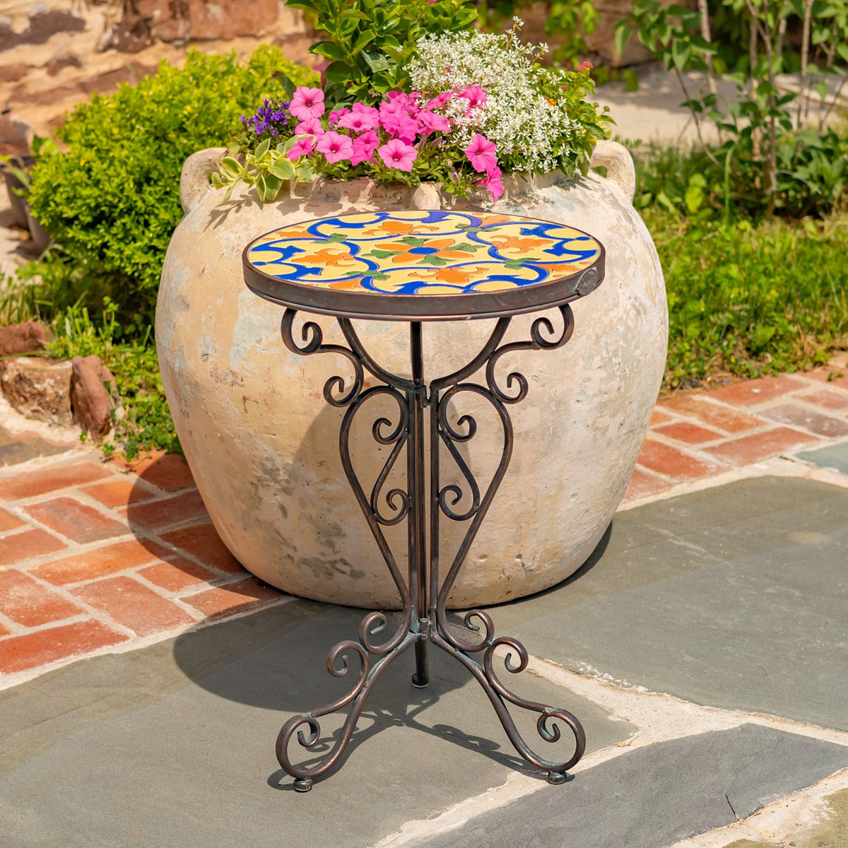 Ornate metal plant stand with a colorful mosaic top beside a large flower-filled planter.