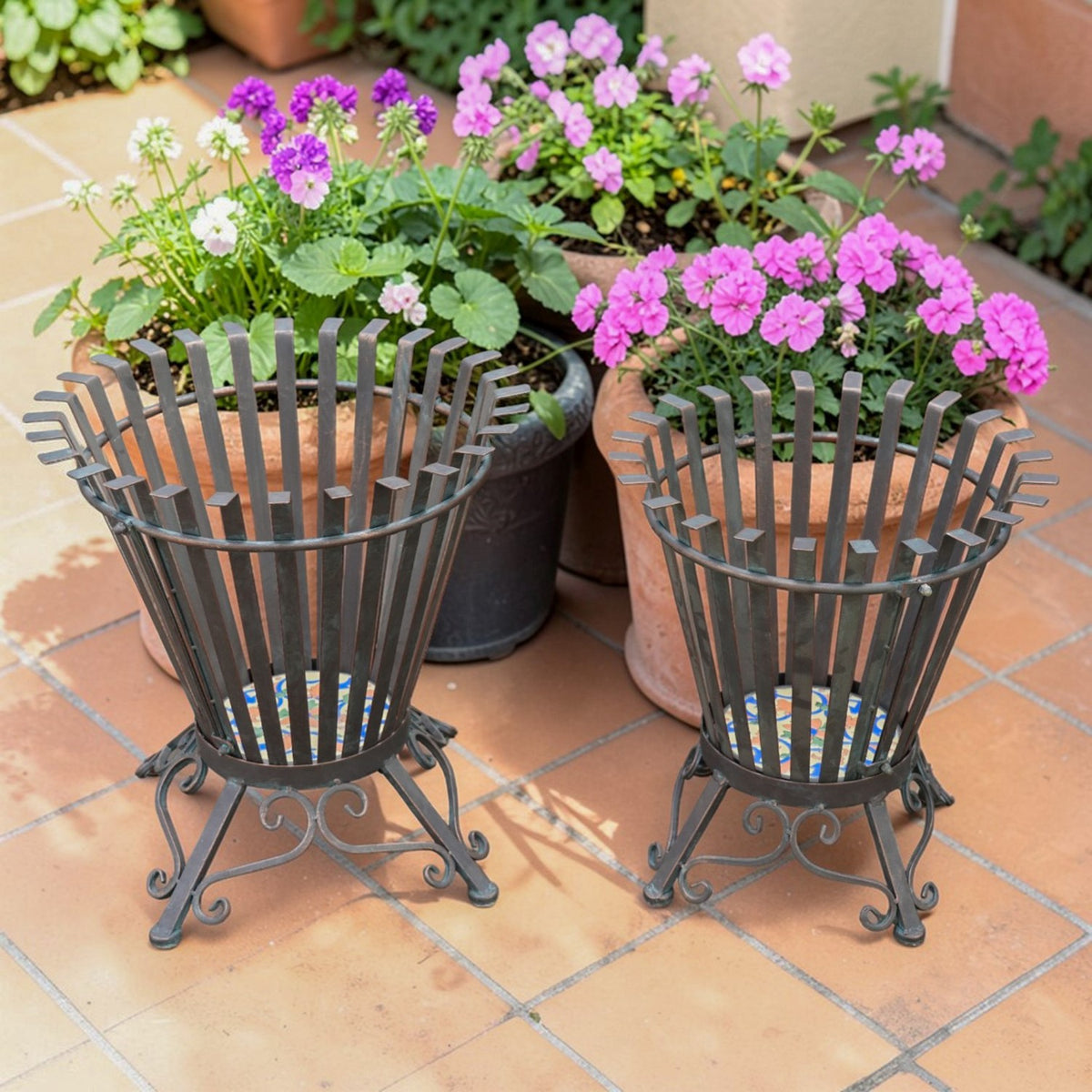 Two dark metal firewood holders with flared slatted sides and mosaic bases on a tiled patio.