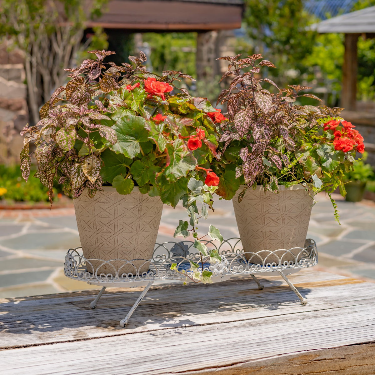 Ornate white metal plant stand holding two flower-filled pots on a wooden outdoor table.