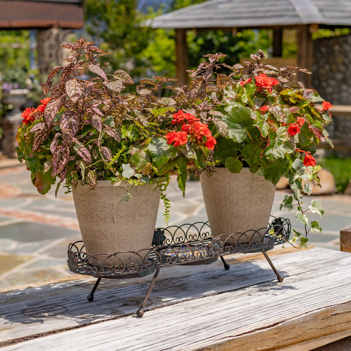 Ornate bronze plant stand holding two flower-filled pots on a sunlit outdoor table.
