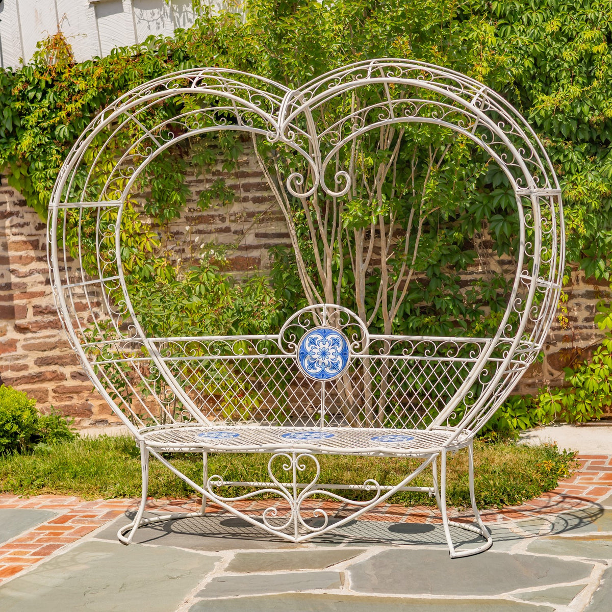 Full view of a white metal bench with a tall heart-shaped back and blue mosaic accents outdoors.