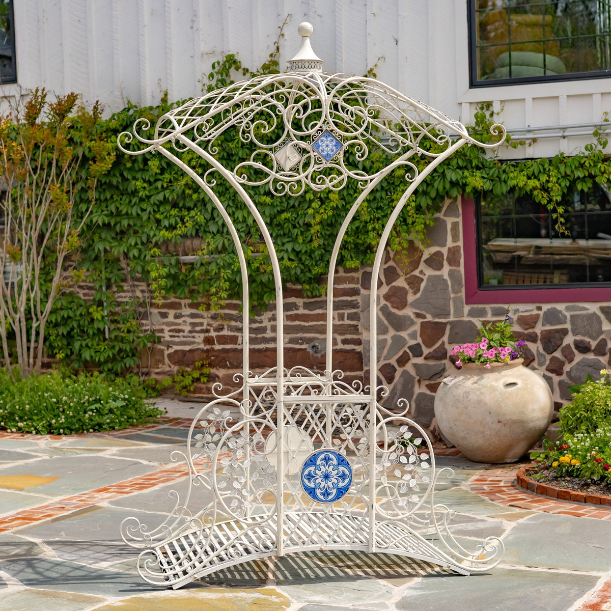 Ornate antique-white metal garden bridge with blue mosaic accents, standing on a stone patio beside a garden wall.