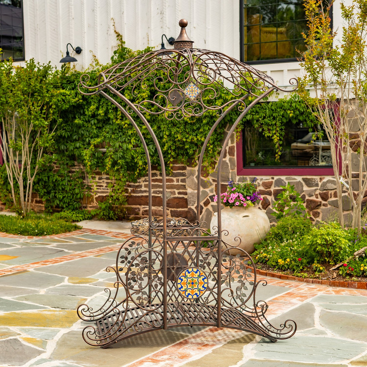 Ornate bronze-colored metal garden bridge with a mosaic accent on a stone patio among greenery.