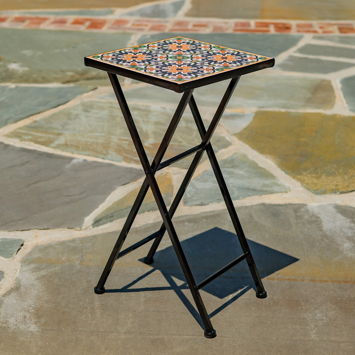 Black folding stand with a square, colorful patterned mosaic top on a stone patio.