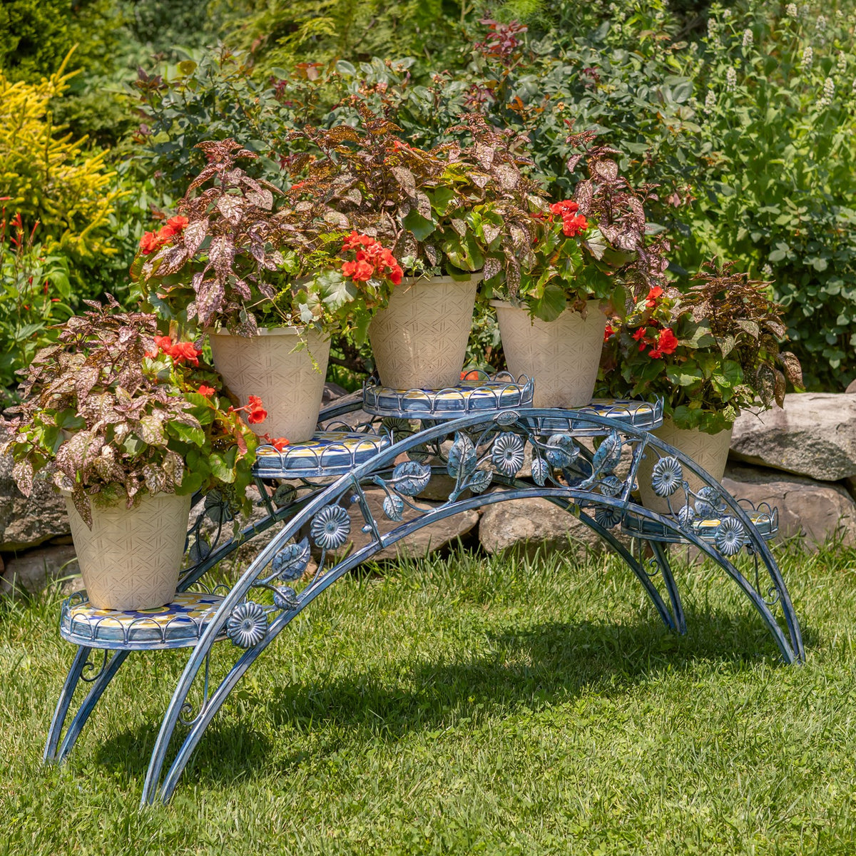Arched blue iron plant stand with patterned mosaic shelves holding potted flowers in a garden.