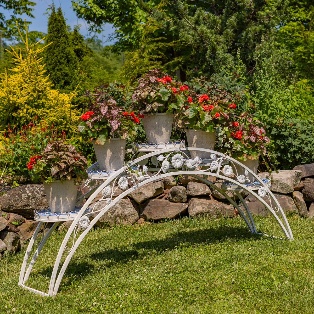 White arched plant stand with blue-and-white mosaic shelves holding flower pots beside a stone wall.