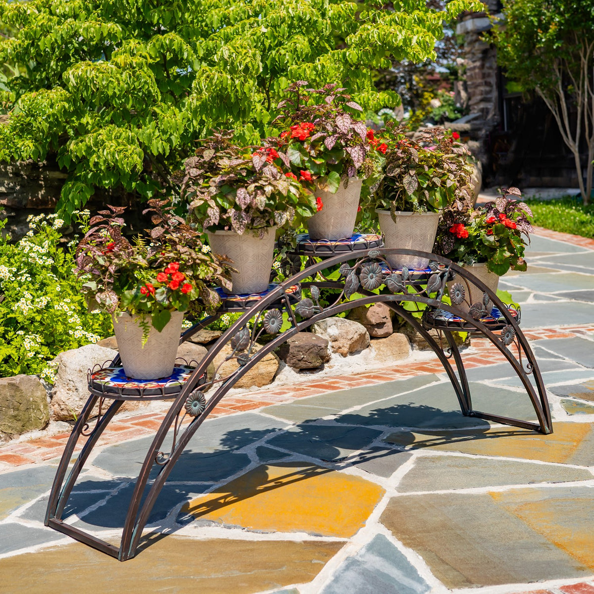 Arched iron plant stand with mosaic shelves displayed outdoors holding potted flowers and greenery.