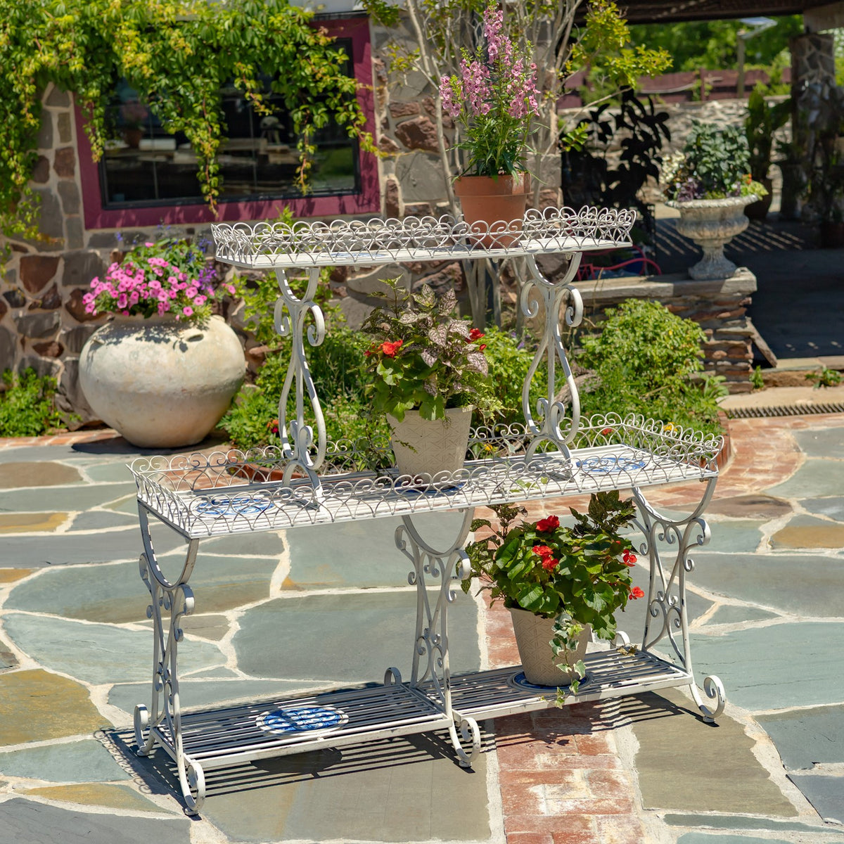 Three-tier white metal display stand with blue mosaic tiles and potted flowers on a stone patio.