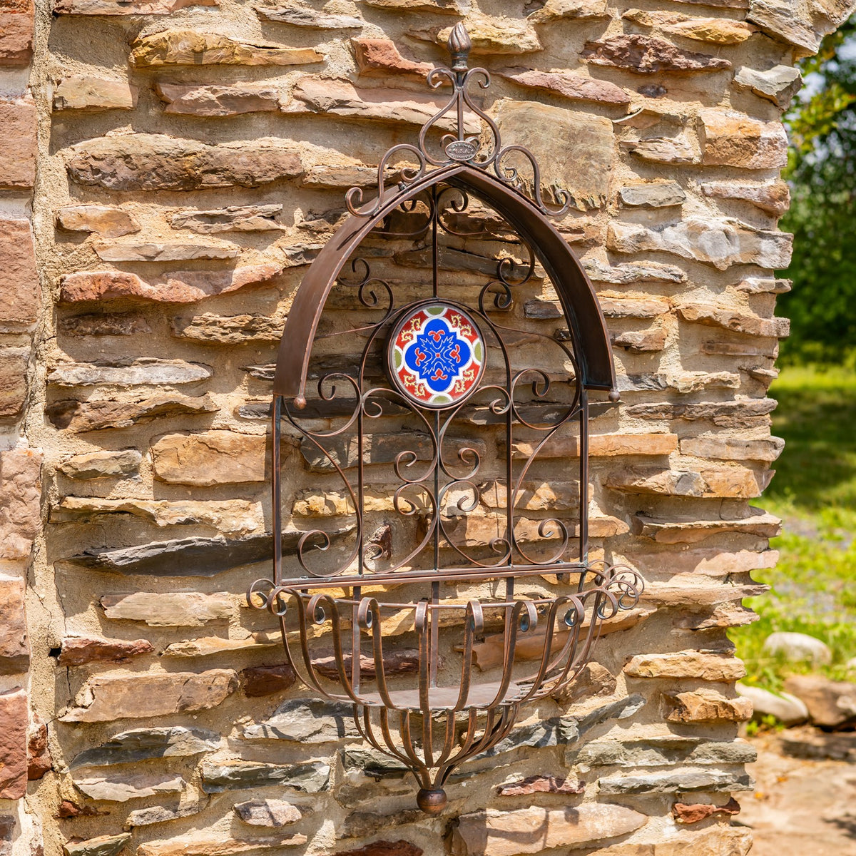 Antique bronze arched wall planter with an ornate iron frame and colorful circular mosaic against a stone wall.