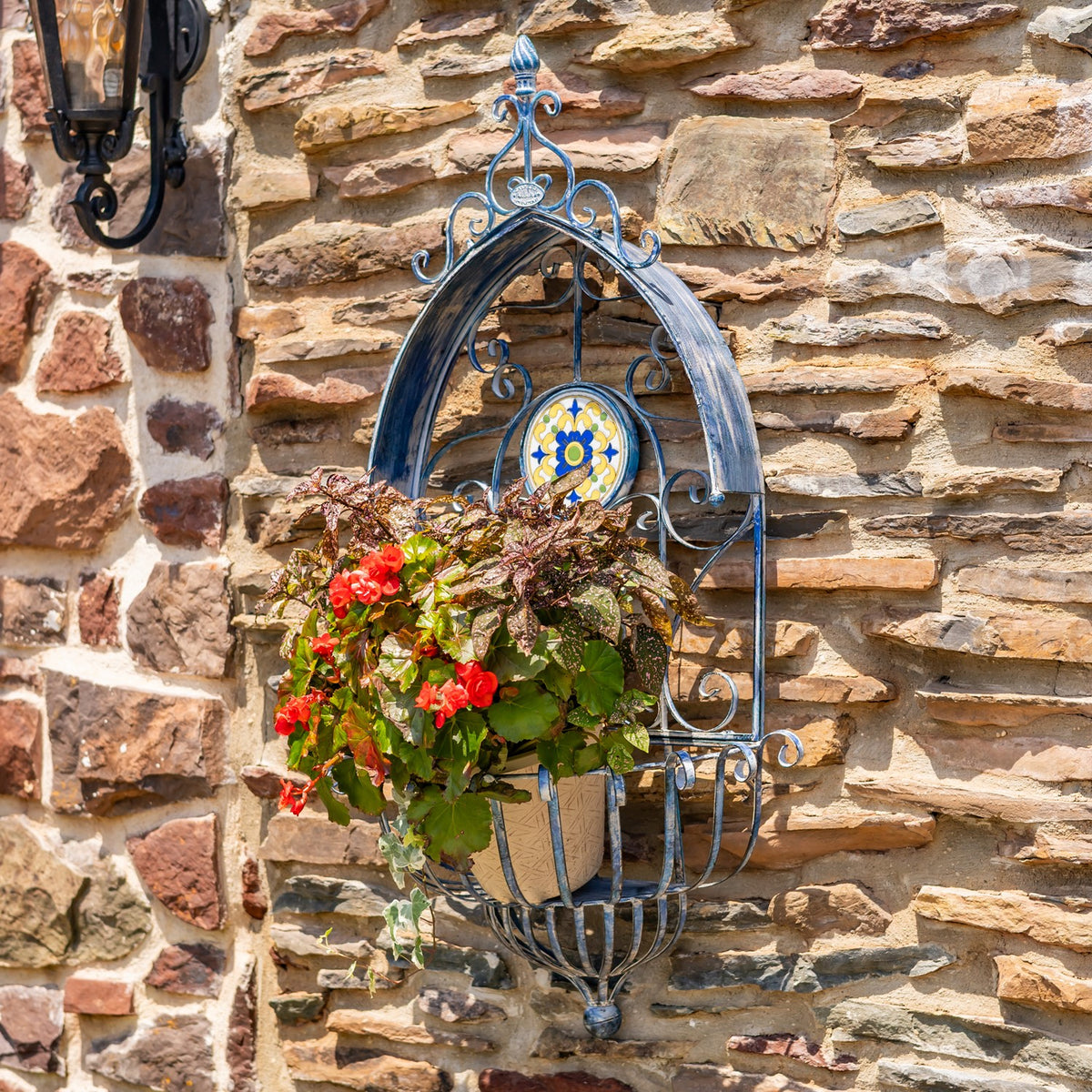 Blue scrollwork wall planter with red flowers and foliage against a rustic stone wall.