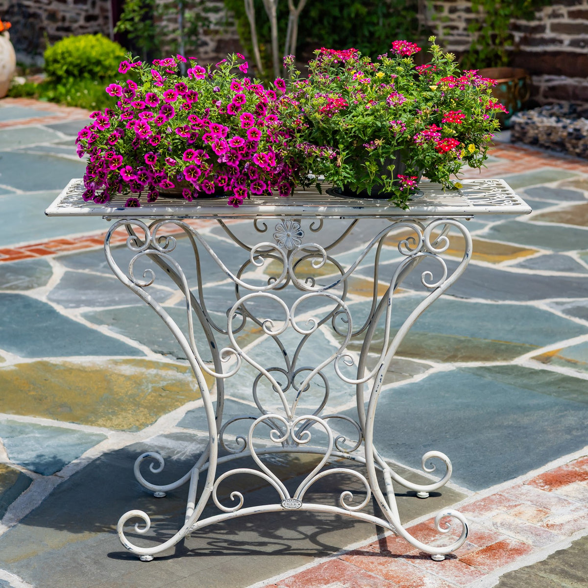 Antique-white scrollwork table with a mosaic tile top, displayed outdoors beneath flower planters.