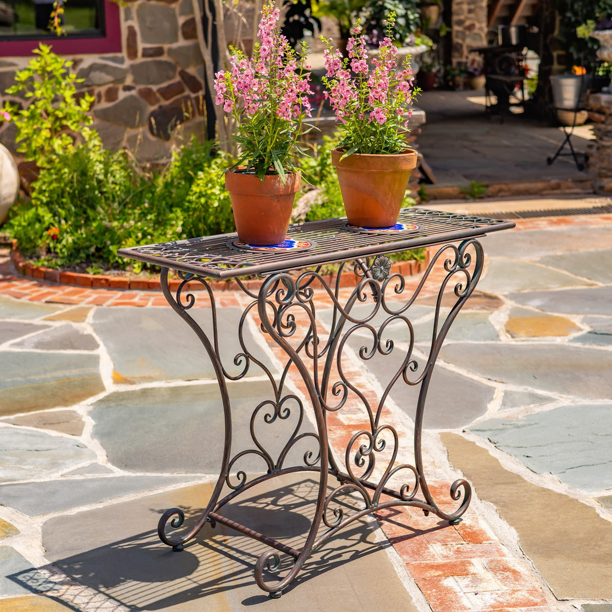Ornate bronze-tone iron table with a mosaic tile top holding two flower pots on a stone patio.